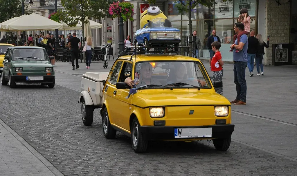 żółty Fiat 126 Maluch z zielonym listkiem ciągnie przyczepkę — nowy kierowca w okresie próbnym na drodze